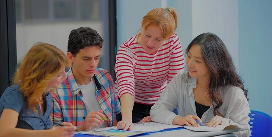 Teacher helping three students at a table with notebooks and stationery.