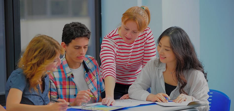 Teacher helping three students at a table with notebooks and stationery.